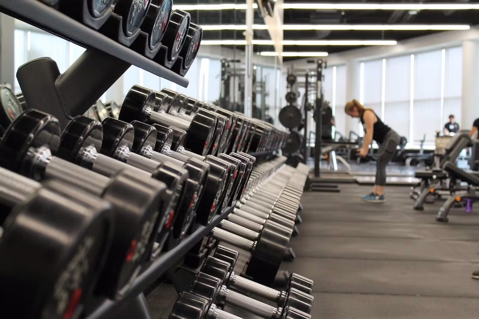 A rack of dumbbells in a gym with a woman lifting weights in front