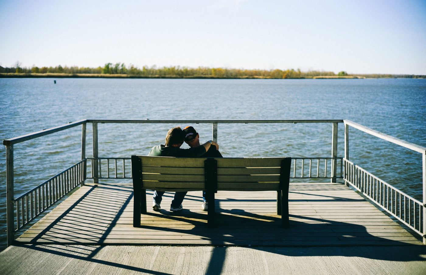 Two men sitting on a public bench in front of a large blue lake.